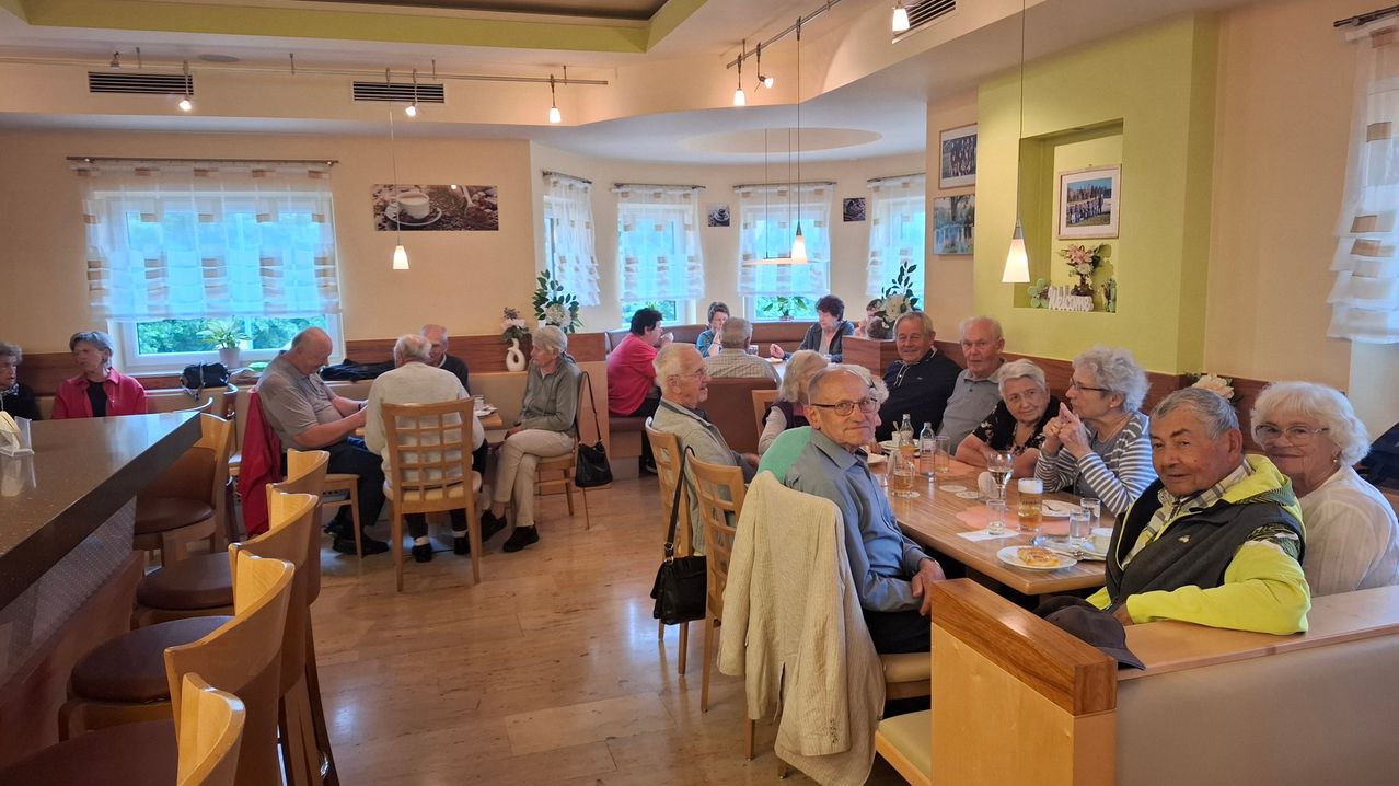Eine Gruppe älterer Menschen sitzt in einem Restaurant. Einige trinken und unterhalten sich. Der Raum hat Holzböden und Wände mit Bilderrahmen.