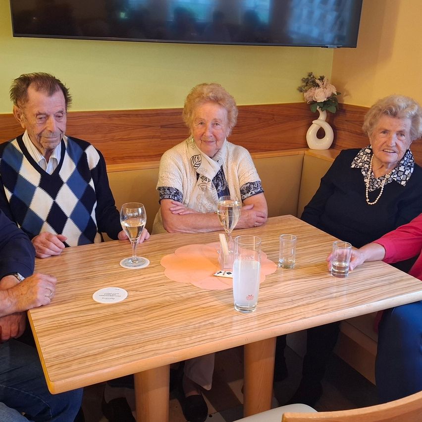 Vier ältere Personen sitzen in einem Restaurant um einen Tisch herum und lächeln. Ein Blumenvase steht auf dem Regal hinter ihnen. Zwei Gläser Wein und drei Gläser Wasser sind auf dem Tisch.