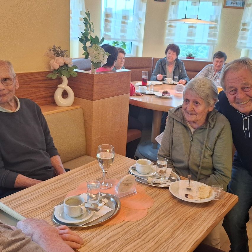 Mehrere ältere Menschen sitzen lächelnd in einem Restaurant. Sie sitzen um einen Tisch mit Tassen, Tellern und einer Vase mit Blumen. Ein weiterer Tisch ist im Hintergrund mit mehr sitzenden Personen.