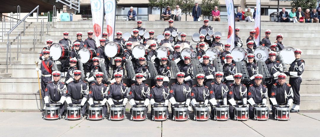 Eine Marschkapelle posiert für ein Foto auf einer Betonfläche. Sie tragen Uniformen, Mützen und halten Trommeln. Im Hintergrund sind Gebäude und Menschen zu sehen.