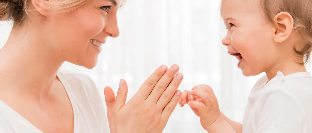 Eine Frau und ein Baby lächeln und spielen miteinander. Die Frau hält die Hände des Babys und das Baby hält die Hände der Frau.