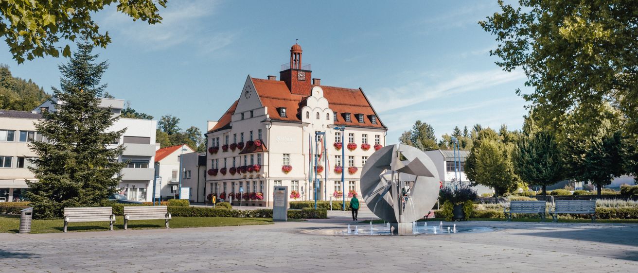 Ein großes weißes Gebäude mit einem roten Dach und mehreren Fenstern mit roten Blumenkästen befindet sich auf einem Stadtplatz mit einem Brunnen und Bänken.