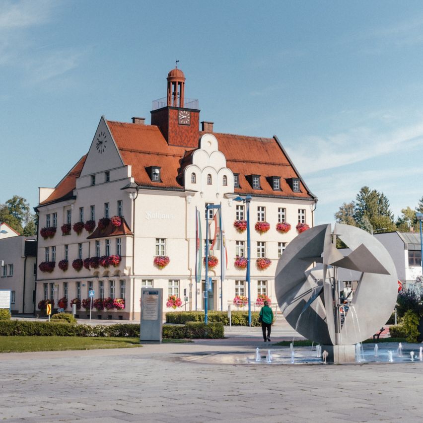 Ein großes weißes Gebäude mit einem roten Dach und mehreren Fenstern mit roten Blumenkästen befindet sich auf einem Stadtplatz mit einem Brunnen und Bänken.