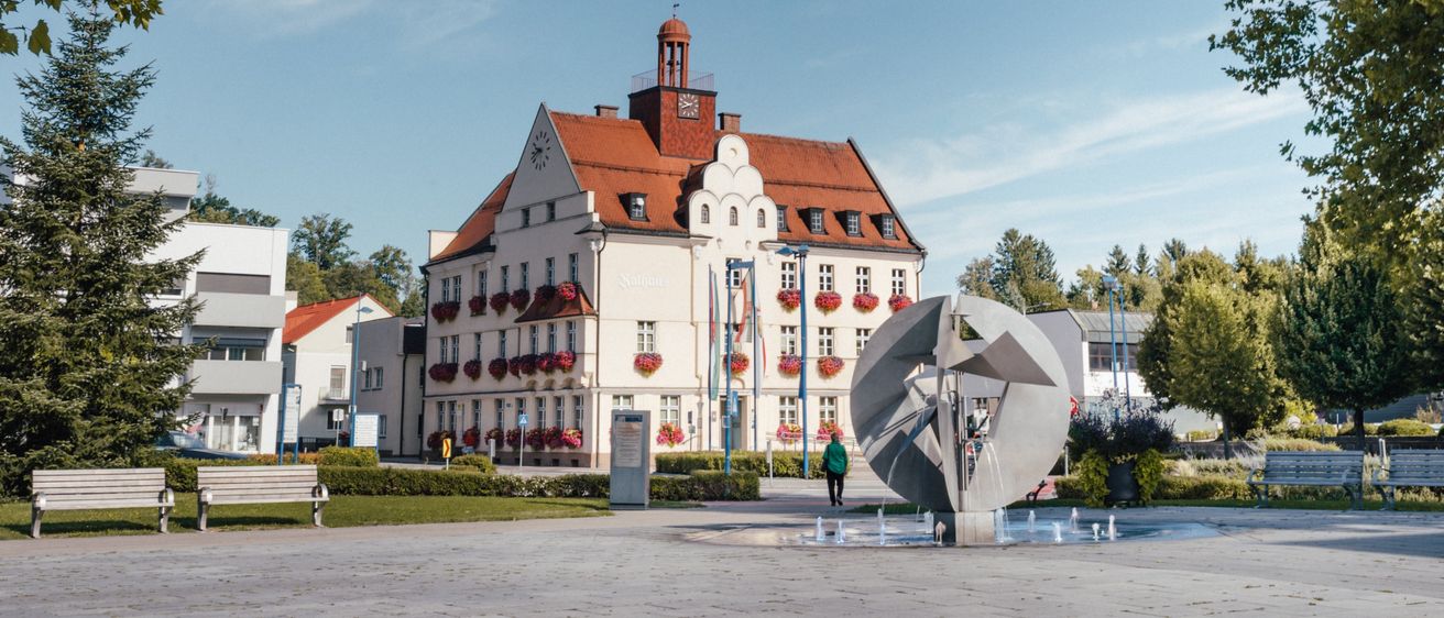 Ein großes weißes Gebäude mit einem roten Dach und mehreren Fenstern mit roten Blumenkästen befindet sich auf einem Stadtplatz mit einem Brunnen und Bänken.