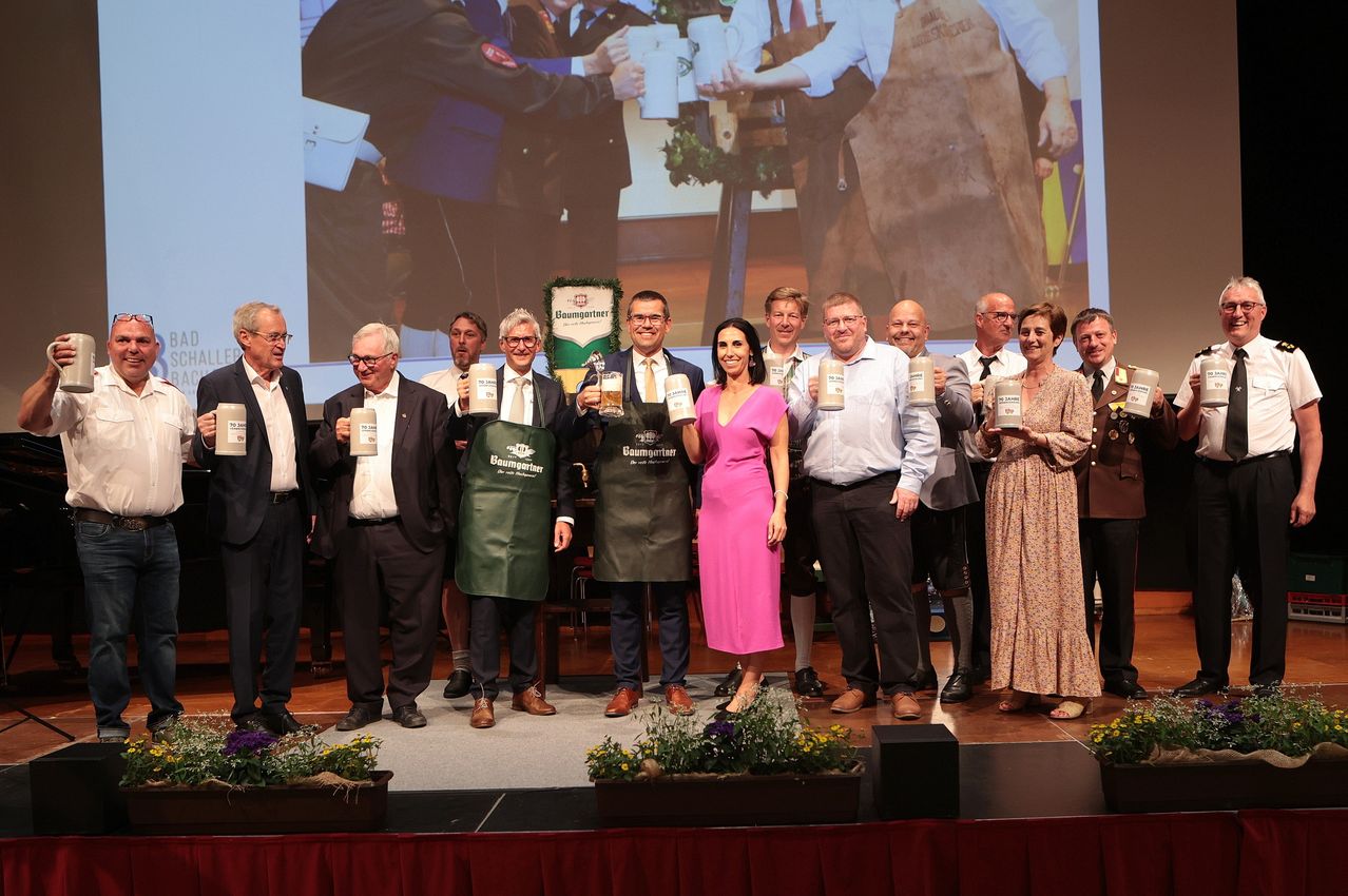 Eine Gruppe von Menschen in formeller Kleidung steht auf einer Bühne und posiert für ein Foto mit einem Hintergrund. Sie halten alle ein Glas mit einem Etikett.