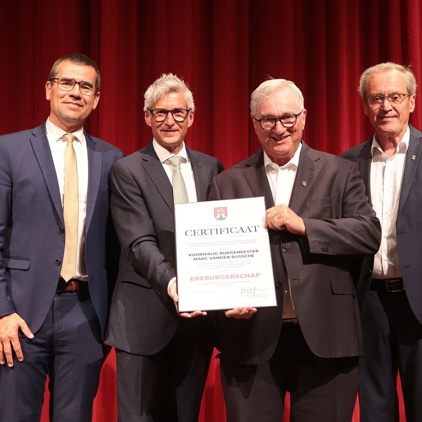 Vier Männer stehen vor einem roten Vorhang. Der Mann in der Mitte hält ein Zertifikat. Der Mann rechts trägt eine Anstecknadel an seiner Jacke.