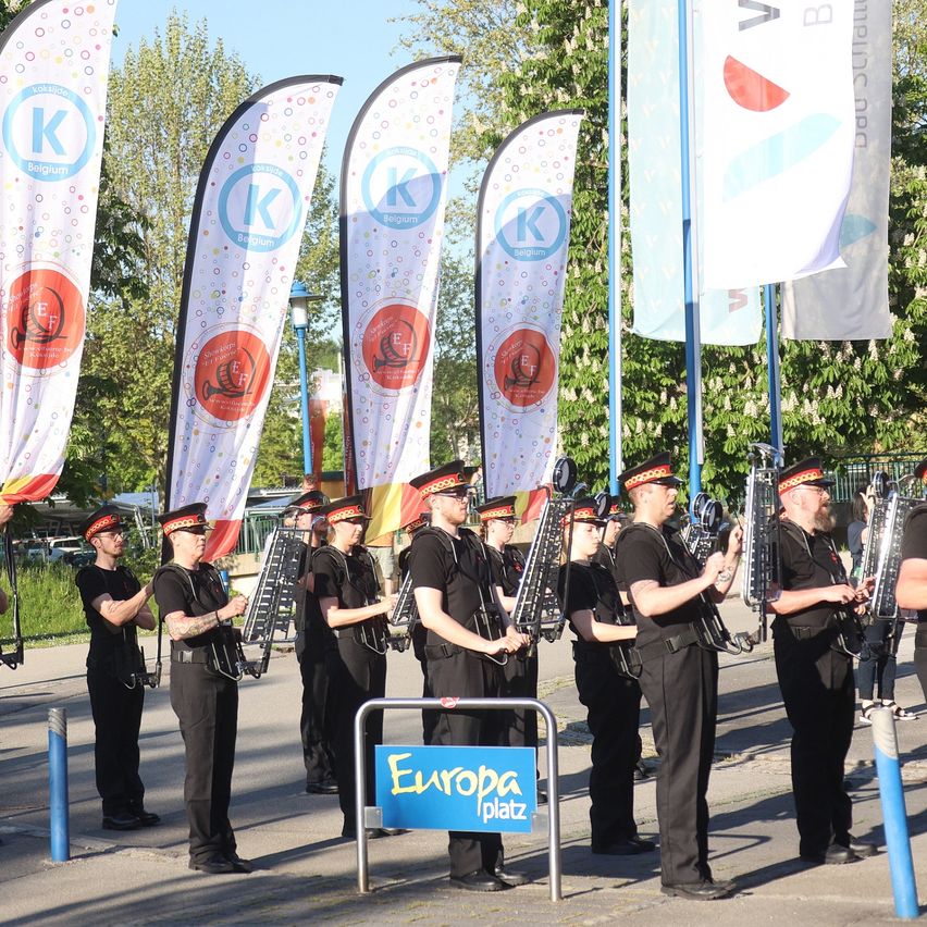 Eine Marschkapelle in schwarzen Uniformen und Mützen, die Musikinstrumente halten, steht vor einem blauen Schild mit der Aufschrift 'Europa Platz', mit verschiedenen Bannern dahinter.