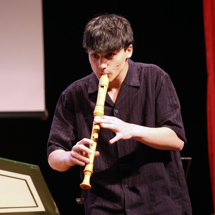 Ein Mann spielt eine Blockflöte bei einer Aufführung. Er steht auf der Bühne mit einem Notenständer hinter ihm und einem roten Vorhang auf der rechten Seite.