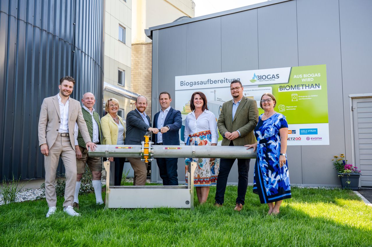 Eine Gruppe von Menschen steht vor einer Biogasanlage und lächelt für ein Foto. Sie stehen auf Gras und im Vordergrund befindet sich eine große Rohrleitung.
