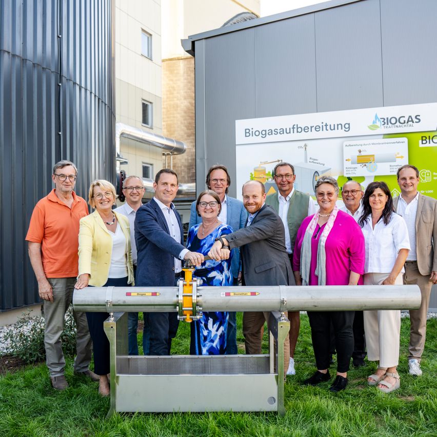 Eine Gruppe von Personen steht um eine Biogasanlage herum und posiert für ein Foto. Die Anlage hat ein Schild mit den Worten 'Biogasaufbereitung' und 'BIOGAS' darauf.