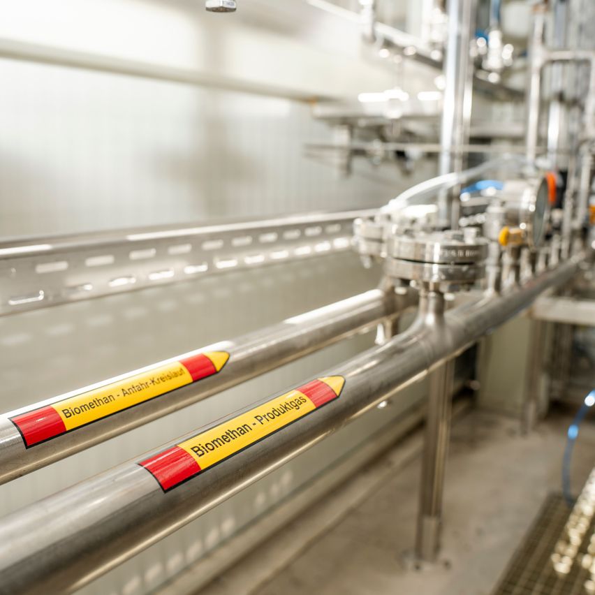 Nahaufnahme von Edelstahlrohren mit den Aufschriften 'Biomethanol - Anfahrt-Kreuslaad' und 'Biomethanol - Produktion's' in einem industriellen Umfeld.