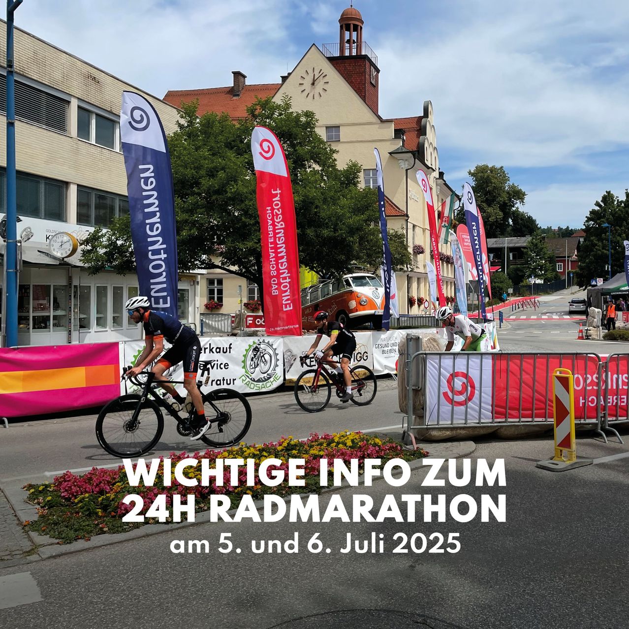 Drei Radfahrer fahren auf einer Straße mit Bannern, die einen 24-Stunden-Radmarathon bewerben, mit einem Gebäude und einem Uhrturm im Hintergrund.