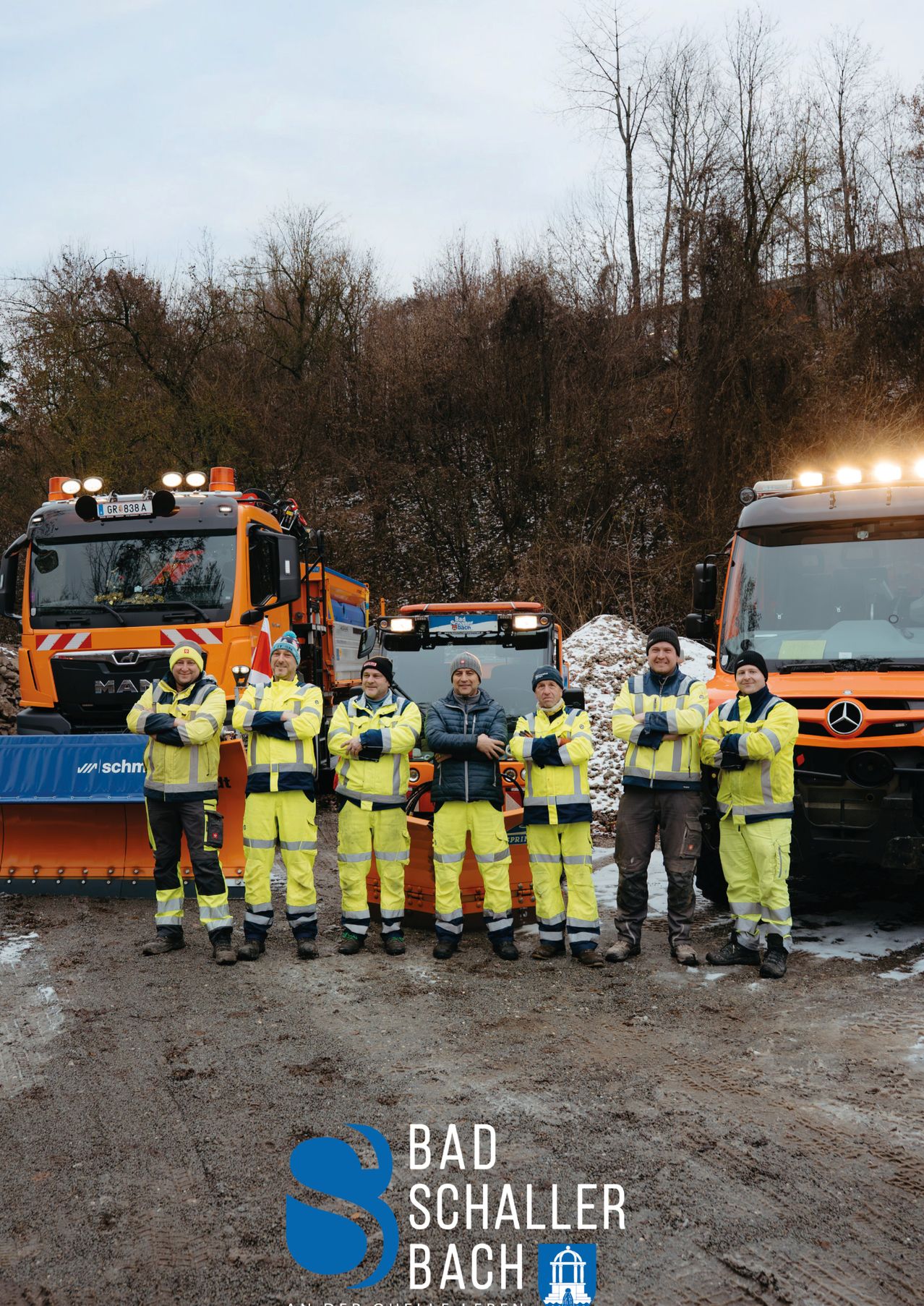 Eine Gruppe von sechs Arbeitern in gelben Schutzanzügen posiert vor drei Schneepflug-Lkws in einer verschneiten Umgebung.