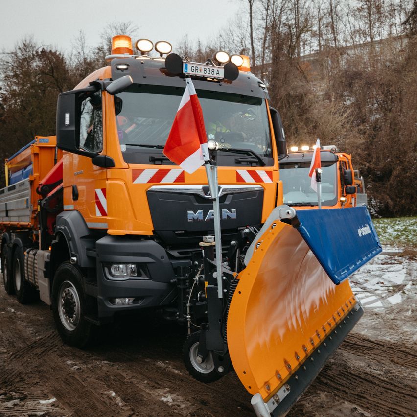 Ein orangefarbener MAN-LKW mit einem Schneepflug steht in einem verschneiten Bereich. Im Hintergrund sind Bäume zu sehen.