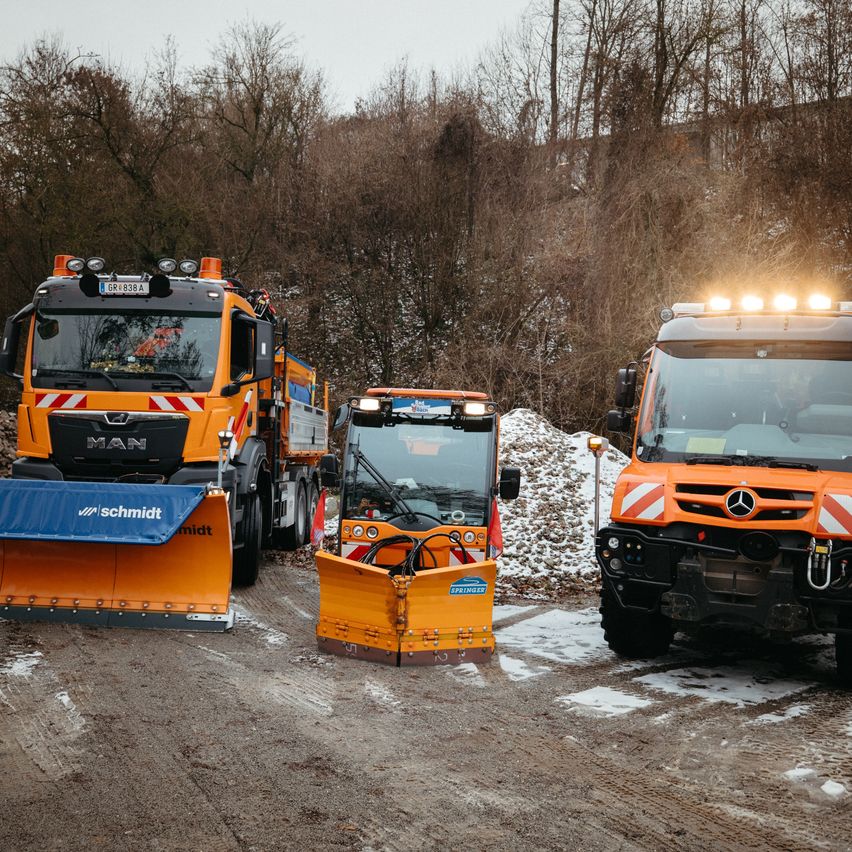 Drei orangefarbene Schneepflug-LKWs sind auf einer verschneiten Straße geparkt. Der vordere LKW hat das Wort MAN auf seiner Front.