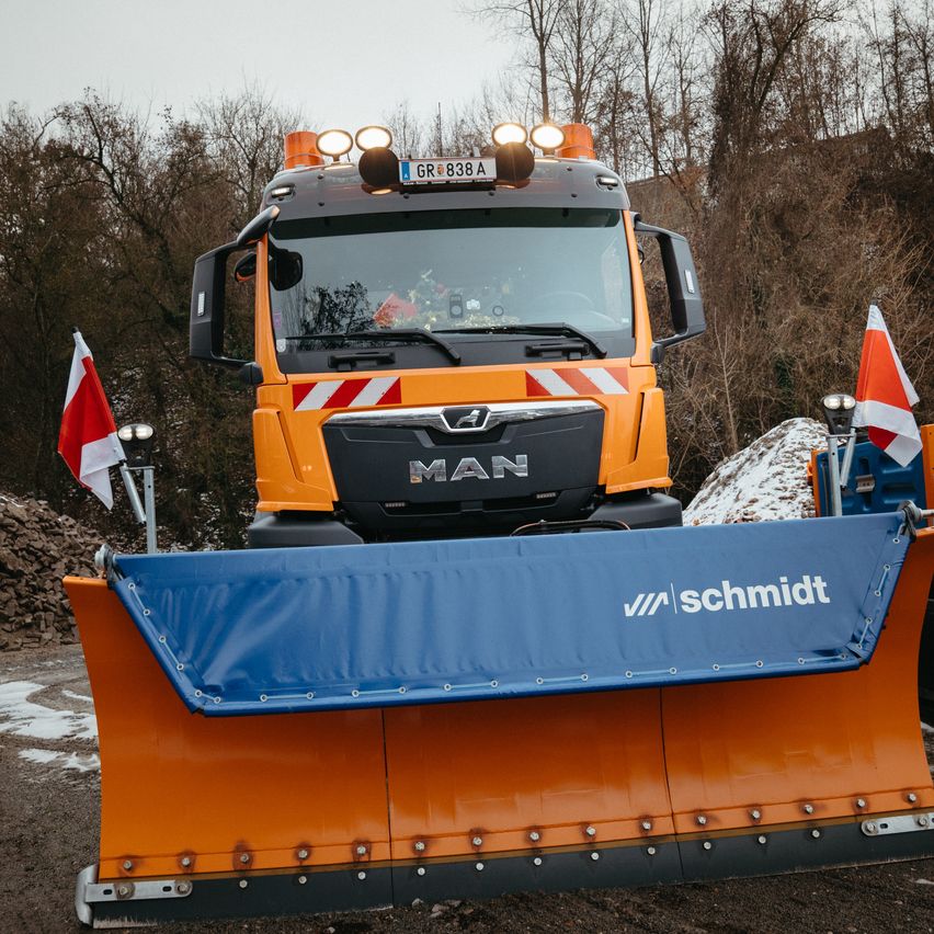 Ein orangefarbener MAN-Lkw mit einem blauen Schneepflug steht auf einer verschneiten Straße. Er hat ein Nummernschild mit der Aufschrift GR 838A.