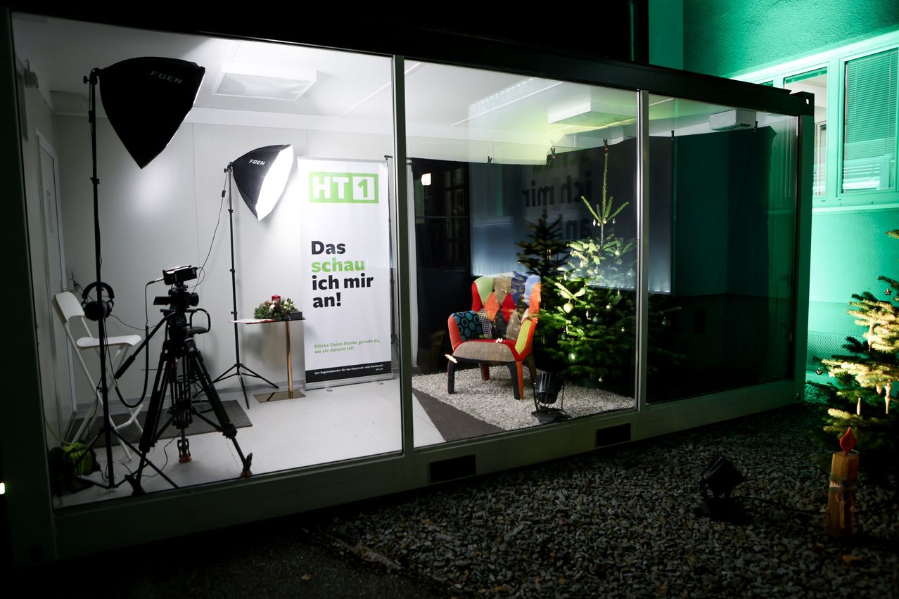 Ein Fotostudio-Setup mit einem Banner, einem Stuhl und einem Weihnachtsbaum in einem Glasraum. Ausrüstung wie eine Kamera auf einem Stativ ist auf der linken Seite sichtbar.