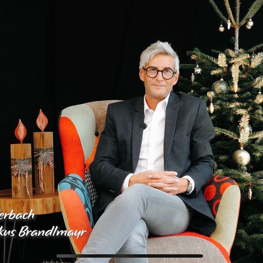 Ein Mann sitzt in einem Stuhl in der Nähe eines geschmückten Weihnachtsbaums. Er trägt einen Anzug und eine Brille. Der Baum hat Ornamente, und zwei Holzkerzen stehen auf einem Tisch hinter ihm. Der Mann hat die Hände gefaltet.