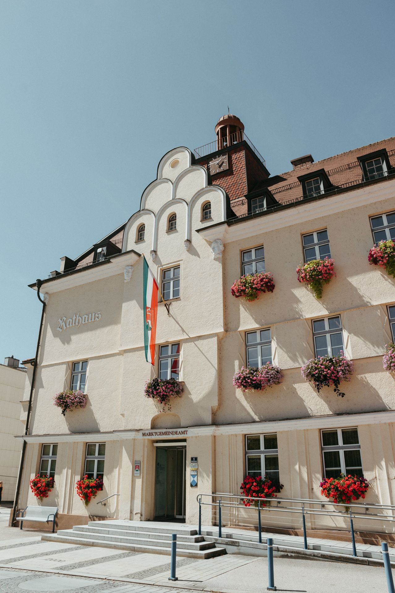 Ein weißes Gebäude mit vielen Fenstern und einem Glockenturm. Eine Flagge weht neben dem Schild, das Rathaus steht. Pinke Blumen schmücken die Fensterbänke.