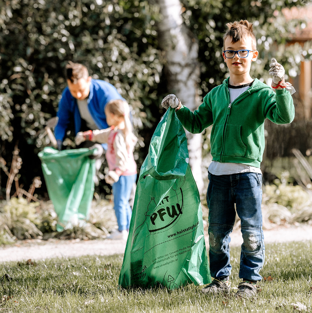 Ein Junge, der einen grünen Beutel hält und Handschuhe trägt, steht auf dem Rasen. Hinter ihm steht ein Mädchen, das einen grünen Beutel hält. Ein Mann in einem blauen Jackett steht neben ihnen. Sie scheinen Müll aufzusammeln.