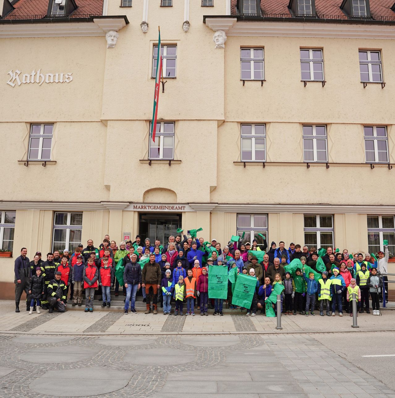 Eine große Gruppe von Menschen, einschließlich Kinder und Erwachsene, steht vor einem Gebäude mit dem Schild Rathaus. Sie halten grüne Banner und scheinen für ein Foto zu posieren.