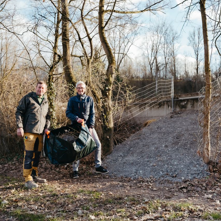 Zwei Männer in Jacken stehen in einem Wald, einer hält einen Beutel. Eine Hecke und ein Weg sind hinter ihnen.