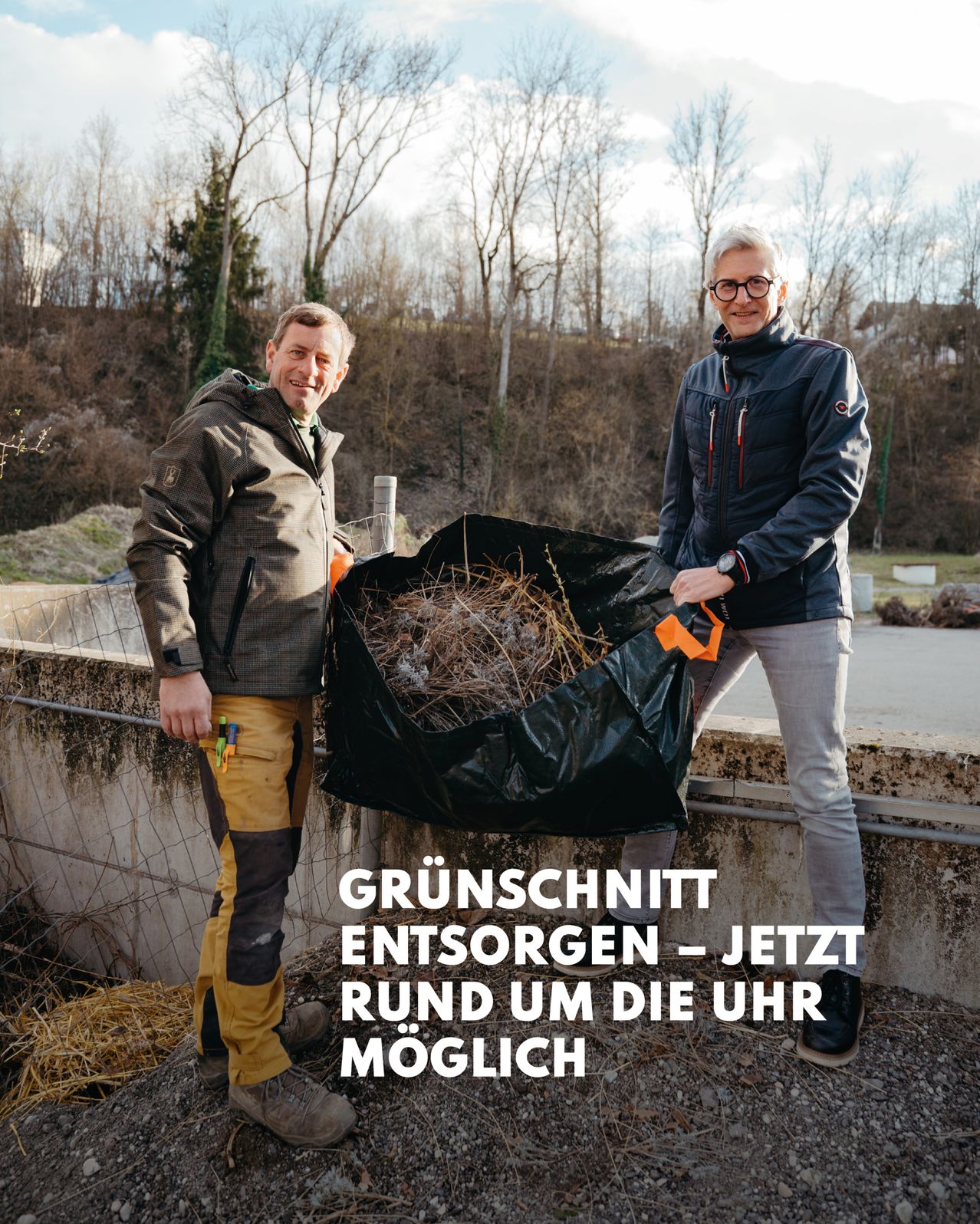 Zwei Männer stehen nebeneinander mit einem großen schwarzen Müllsack. Der Mann auf der linken Seite hält eine Schaufel und der Mann auf der rechten Seite hält den Sack. Dahinter befindet sich eine Wand und ein Grasbereich.