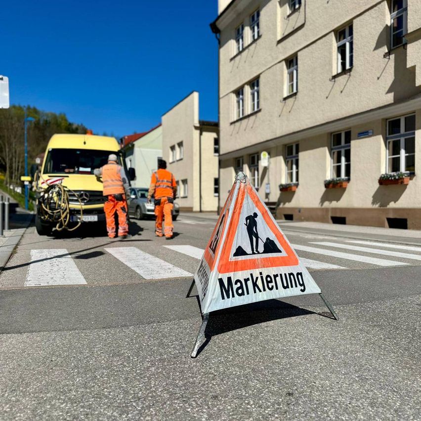 Zwei Arbeiter in orangefarbenen Warnwesten überqueren eine Straße mit einem Straßenbauzeichen. Ein gelber Van ist in der Nähe geparkt. Dahinter befindet sich ein Gebäude mit mehreren Fenstern und einer Straßenlaterne.