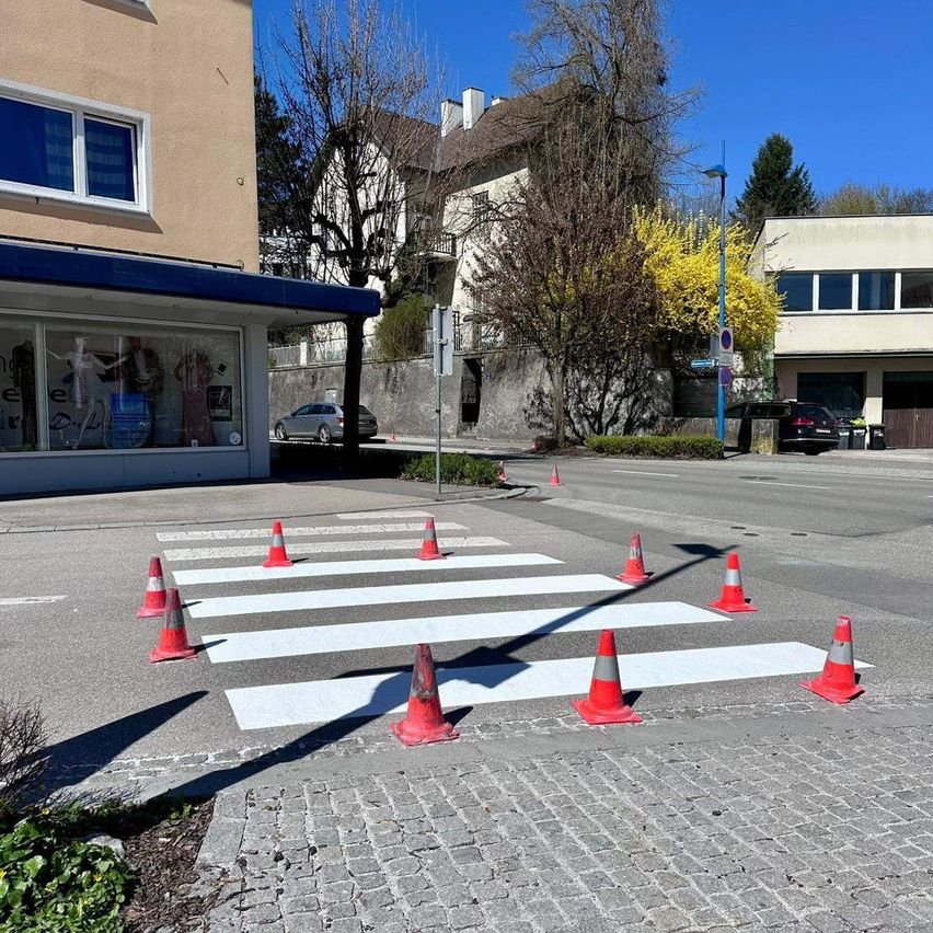 Eine Straße mit einem Zebrastreifen, der durch weiße Linien und Verkehrskegel gekennzeichnet ist, mit Gebäuden und Autos im Hintergrund.