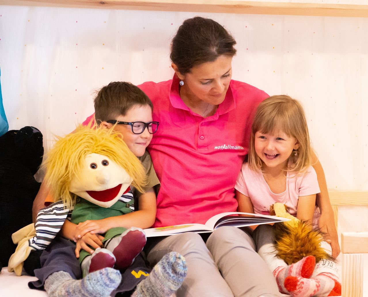 Eine Frau mit einem rosa Shirt liest zwei Kindern ein Buch vor. Ein Junge hält eine Puppe, und ein Mädchen sitzt neben ihm.