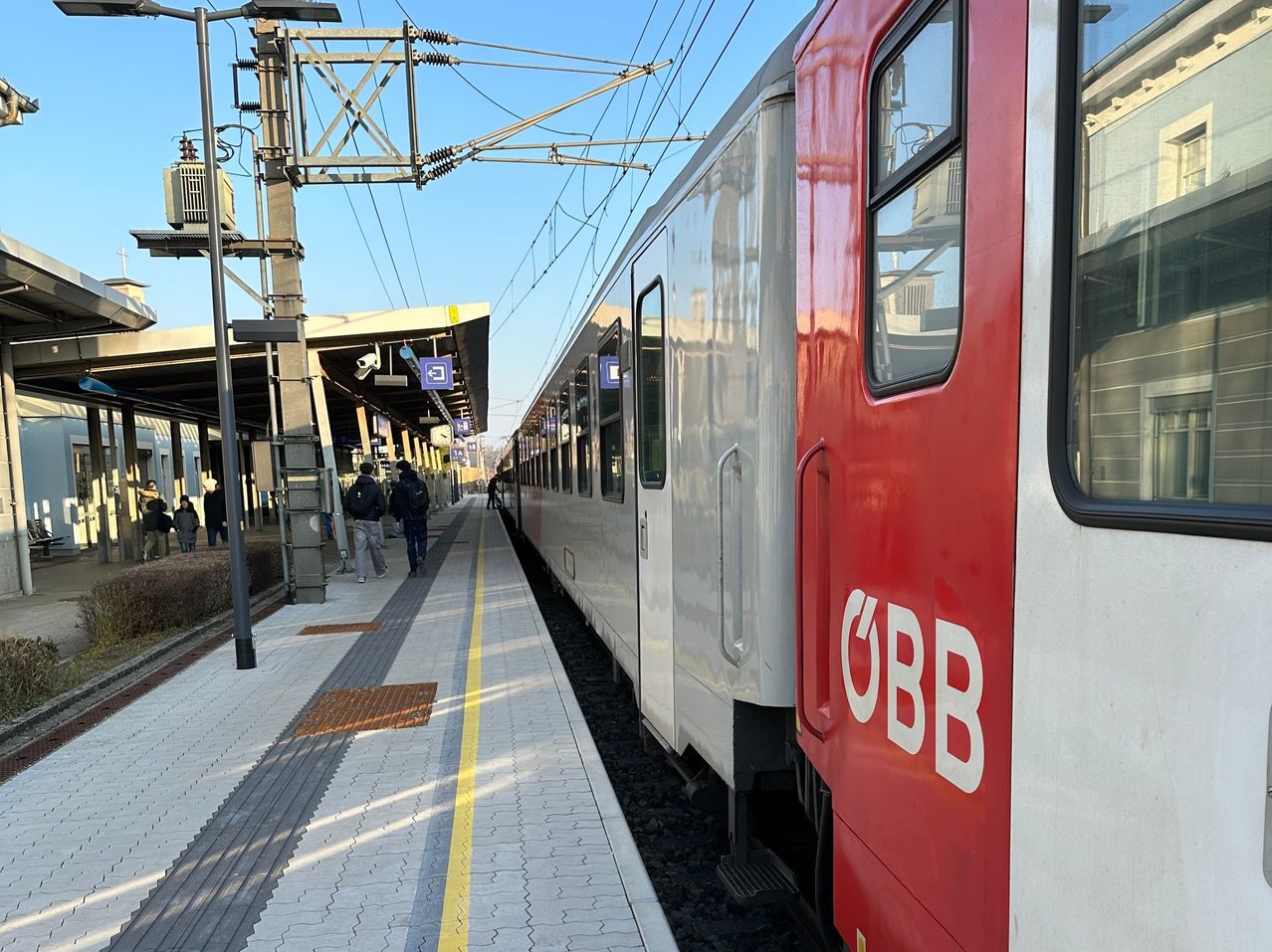 Ein rot-weißer Zug mit der Aufschrift OBB steht auf einem Bahnsteig, in der Nähe gehen Menschen vorbei.