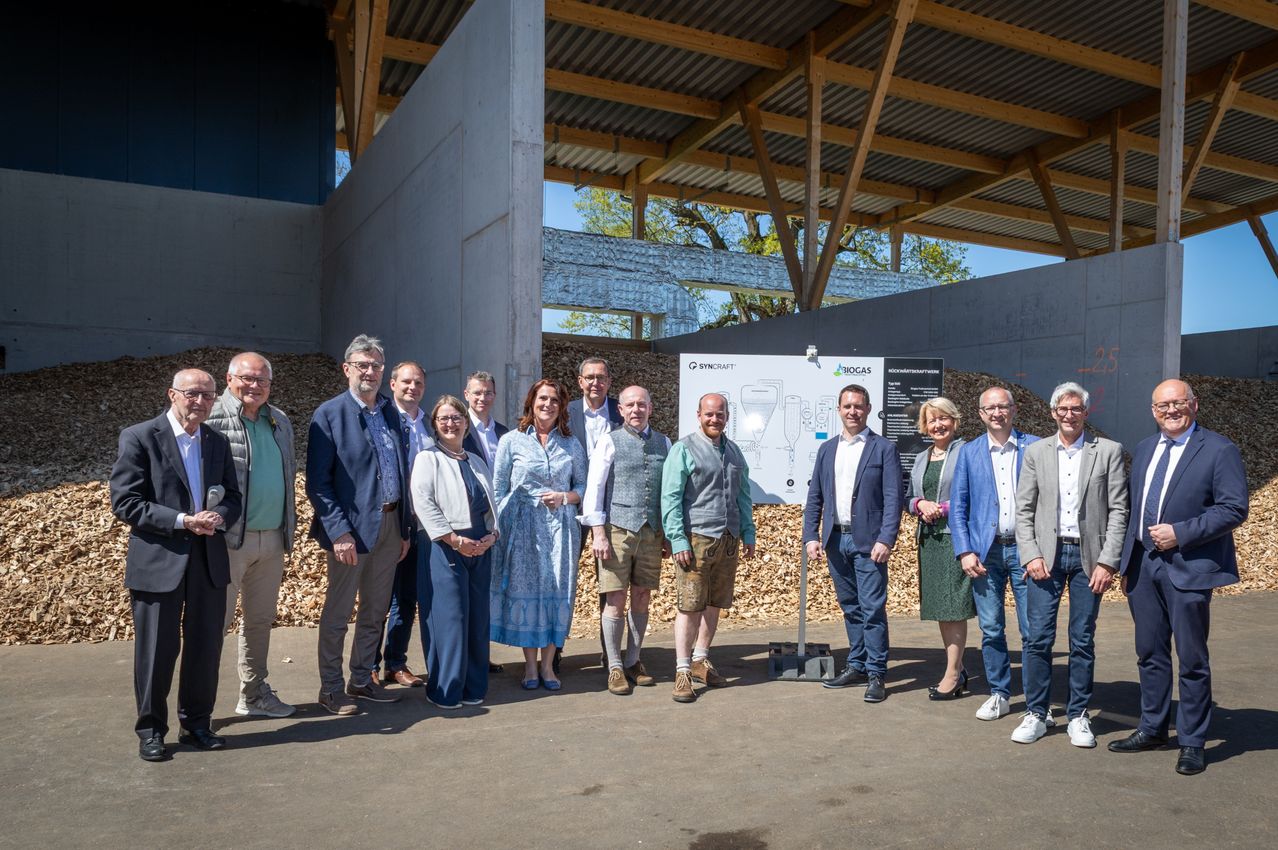 Eine Gruppe von Personen steht vor einem großen weißen Schild mit dem Wort Biogas, mit einer Holzstruktur im Hintergrund.