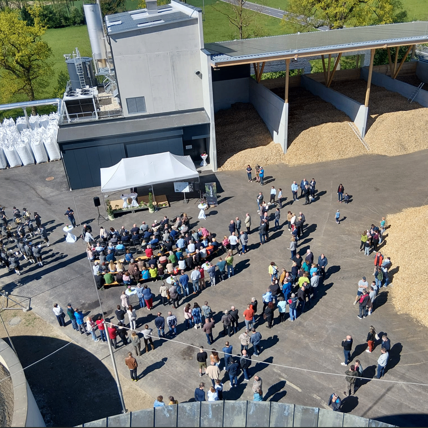 Ein Luftbild einer Industrieanlage mit zahlreichen Menschen, die sich in einem Freiluftbereich versammelt haben. Ein weißes Zelt ist aufgestellt, und es stehen Leute in der Nähe. Im Hintergrund sind große Haufen von Getreide zu sehen.