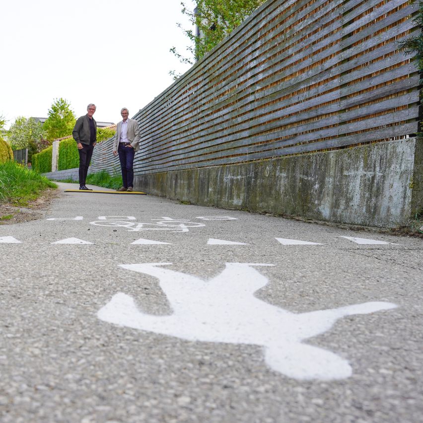Zwei Männer stehen auf einem Weg mit einem Fußgängerübergangsschild und einer Silhouette. Dahinter steht ein Holzzaun.