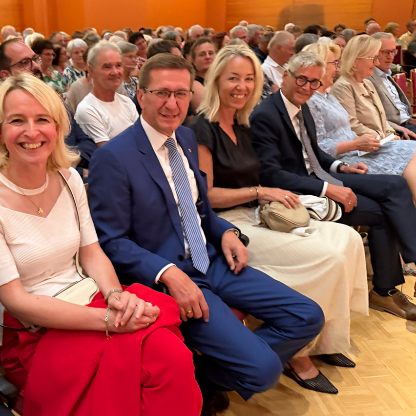 Eine Gruppe von Menschen sitzt in einem Saal, ein Mann in Anzug lächelt mit einer Frau vor ihm.