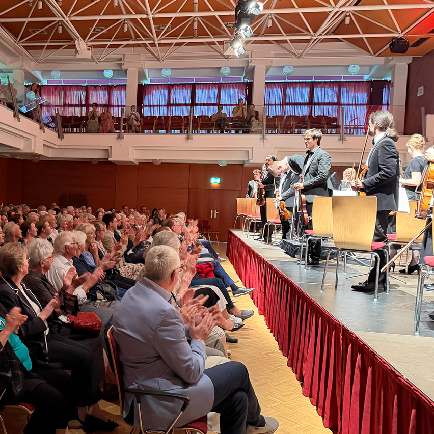 Ein Publikum klatscht während einer klassischen Musikaufführung in einem großen Saal. Die Musiker, einer mit einer Geige, stehen auf der Bühne mit roten Vorhängen dahinter.