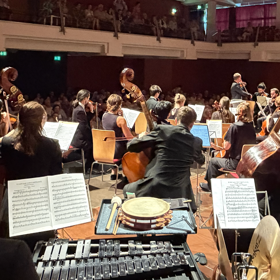 Ein Symphonieorchester spielt auf der Bühne mit Geigern, Cellisten und Bläsern. Das Publikum sitzt in der Galerie und beobachtet. Die Musiker halten offene Notenblätter in den Händen, mit einem Schlagzeug und Xylophon davor.