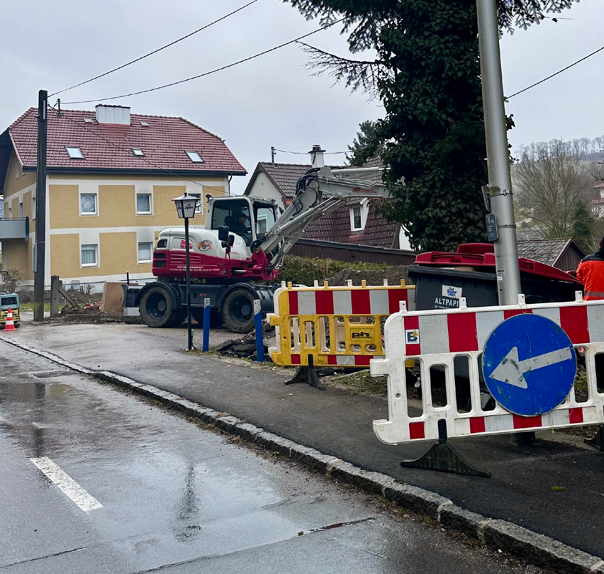 Ein roter und weißer Bagger steht am Straßenrand mit Bauabsperrungen in der Nähe. Ein Haus mit rotem Dach ist hinter dem Bagger sichtbar.