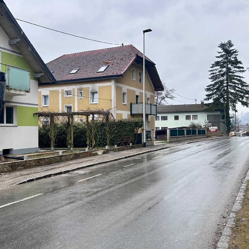 Eine nasse Straße gesäumt von Häusern. Ein gelbes Gebäude mit rotem Dach und Balkon. Grünes und weißes Gebäude auf der linken Seite. Bäume und Pflanzen an der Seite.