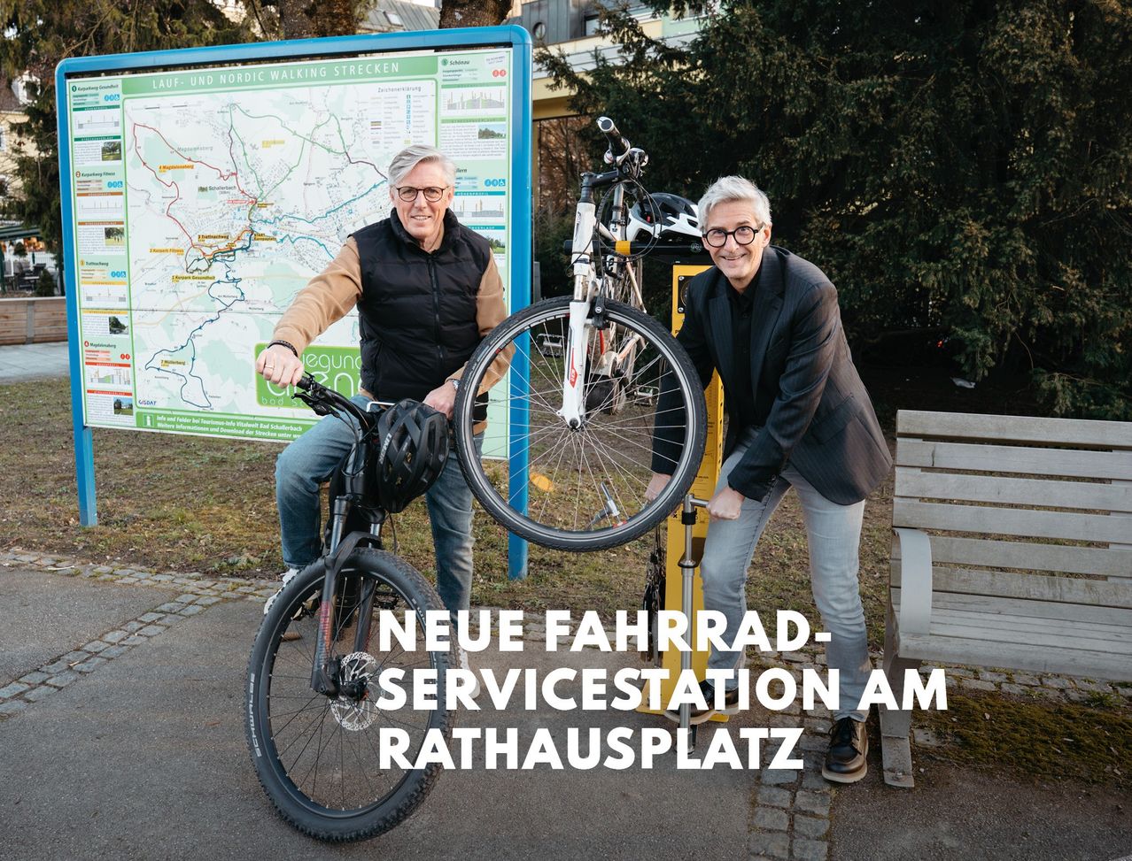 Zwei Männer posieren mit Fahrrädern neben einer neuen Fahrradservicestation am Rathausplatz, mit einer Karte im Hintergrund.