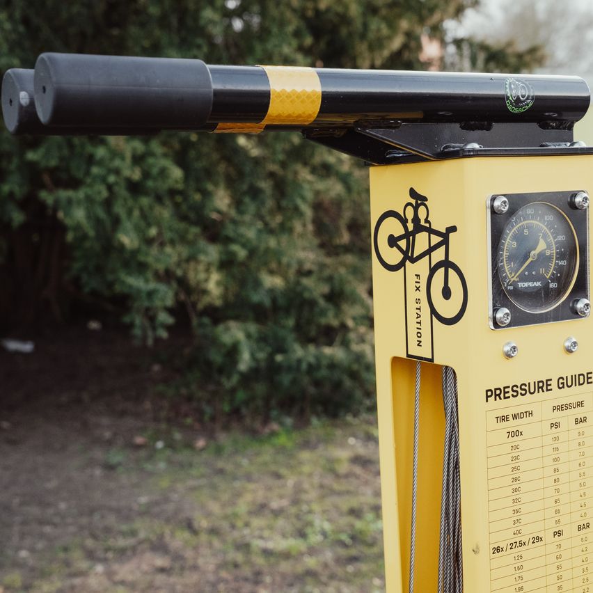 Eine gelbe Fahrrad-Reparaturstation mit einem Druckführungsdiagramm und einem Fahrrad-Symbol. Die Anleitung listet Reifenbreiten und entsprechende PSI- und BAR-Drücke auf.