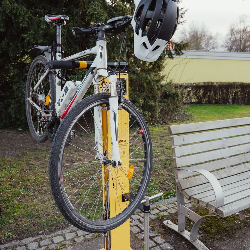 Ein weißes Fahrrad mit schwarzem Helm ist an einem gelben Fahrradständer neben einer Bank auf einer Rasenfläche befestigt.