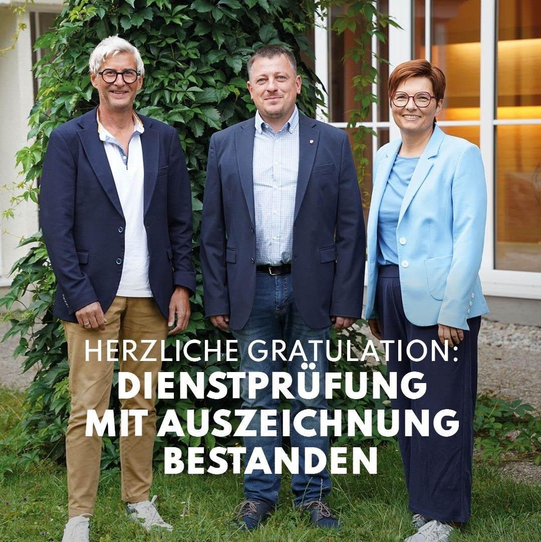 Drei Personen in formeller Kleidung stehen zusammen auf einem Rasen und lächeln für ein Foto. Der Mann links trägt eine Brille und einen Gürtel, der Mann in der Mitte trägt einen Gürtel und eine Uhr, und die Frau rechts trägt eine Brille und einen Ring. Hinter ihnen ist ein Busch und ein Gebäude mit Glasfenstern zu sehen. Der Textüberlagerung lautet 'HERZLICHE GRATULATION: DIENSTPRUFUNG MIT AUSZEICHNUNG BESTANDEN'.