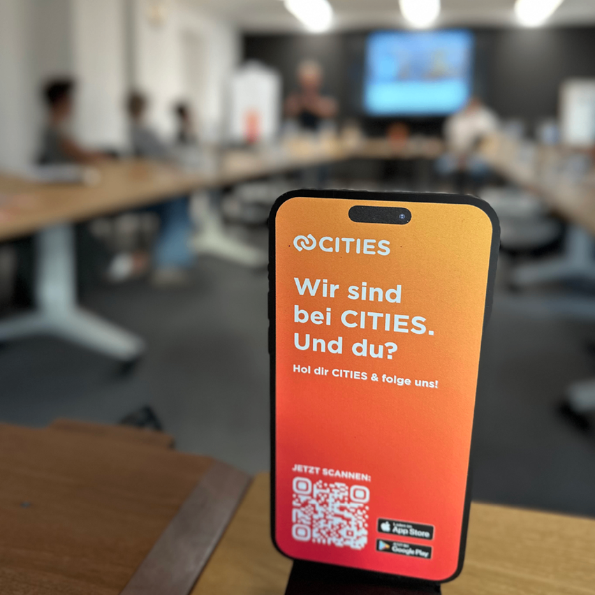 Ein Smartphone zeigt eine Werbung für CITIES mit einem QR-Code, auf einem Holztisch in einem verschwommenen Hintergrund eines Klassenzimmers.
