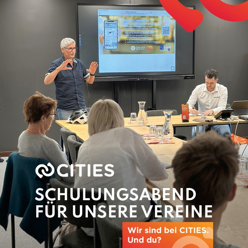 Eine Gruppe von Menschen in einem Raum mit einer Projektionsleinwand, die eine Präsentation zeigt. Ein Mann spricht, während andere zuhören. Der Text auf dem Bildschirm lautet 'Veranstaltungen Immer dabei, mit CITIES!'.