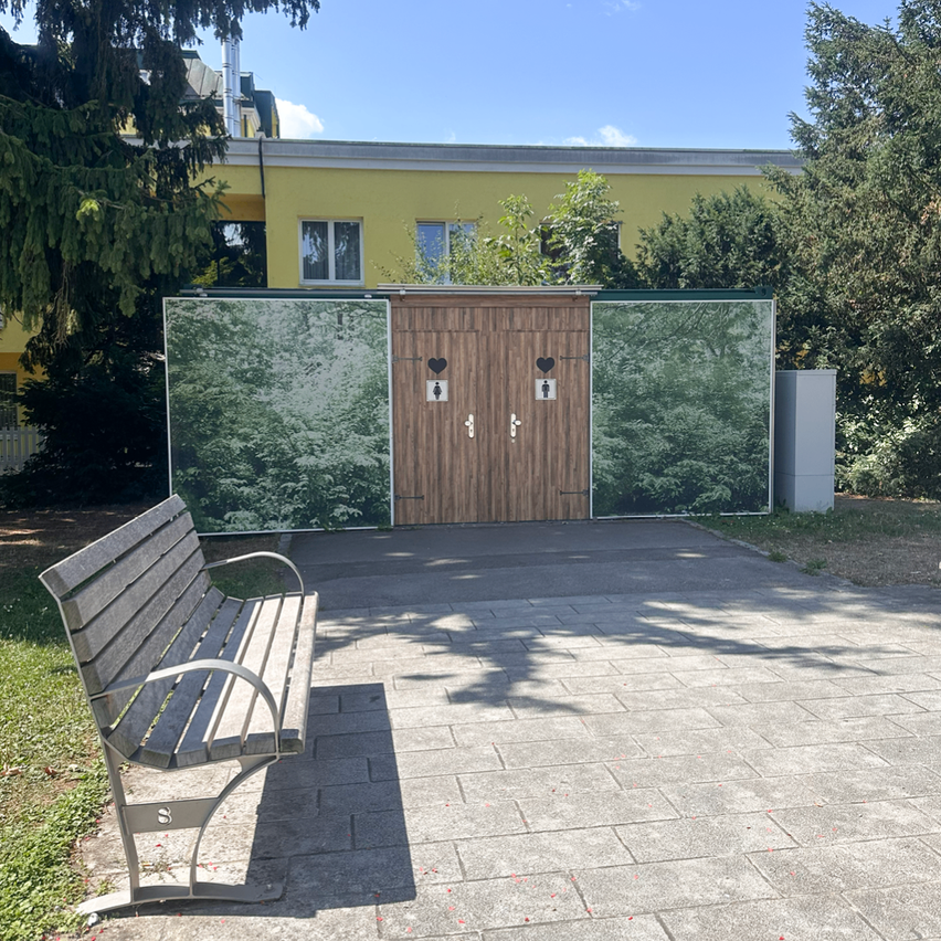 Eine hölzerne Tür mit zwei Herzen und Toilettensymbolen steht vor einem gelben Gebäude, mit einer Bank in der Nähe.