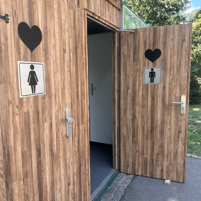 Eine hölzerne Toilettenkabine mit zwei Türen, eine mit einem weiblichen Symbol und die andere mit einem männlichen Symbol, beide mit einem schwarzen Herz darüber.