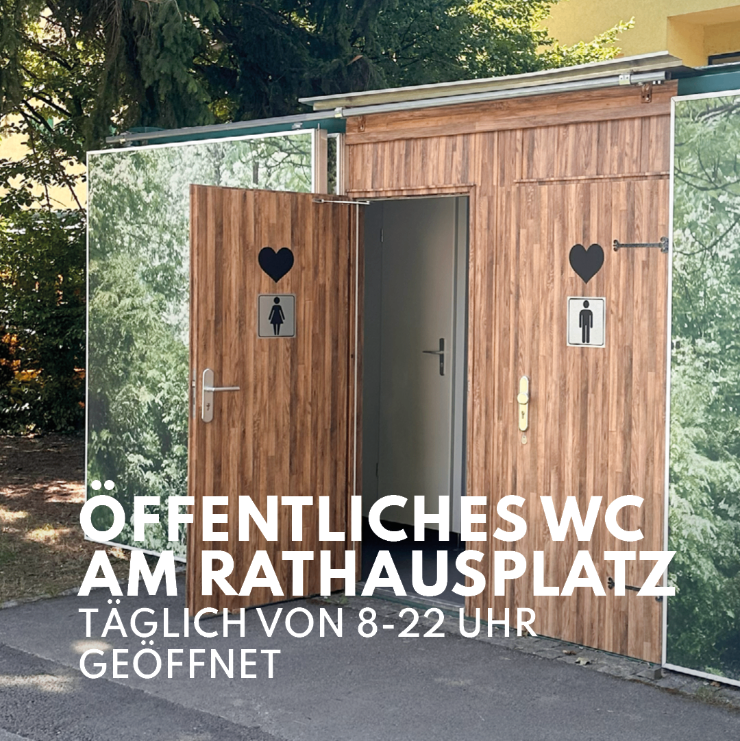 Eine öffentliche Toilettenkabine aus Holz mit einer Glastür und einem Herzsymbol auf der Tür. Die Kabine ist geöffnet, und der Text auf der Tür lautet 'Öffentliches WC am Rathausplatz' und 'Geöffnet'.