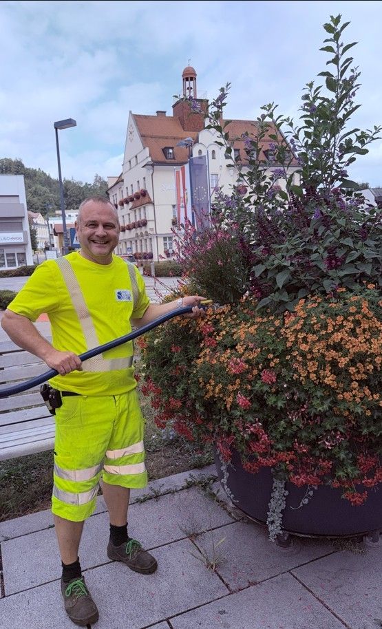 Ein Mann in einem gelben Sicherheitsanzug sprüht Wasser auf einen großen Blumenkasten mit bunten Blumen vor einem Gebäude.