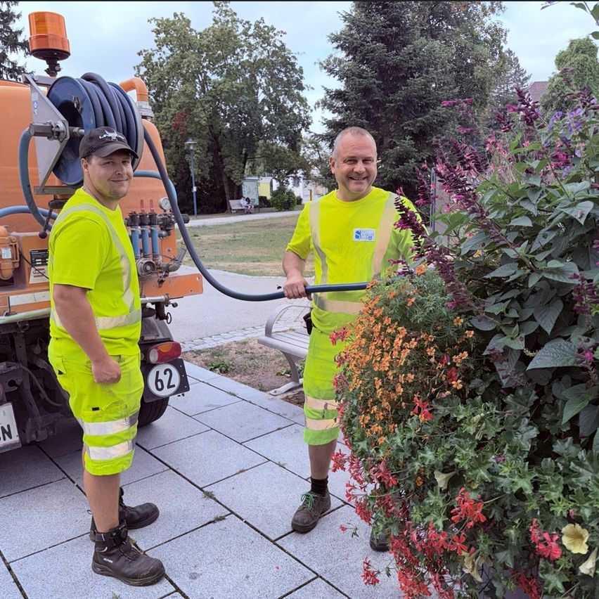 Zwei Männer in neongelben Warnschutzanzügen stehen neben einem Blumenbeet, einer hält eine Gartenschlauch. Sie lächeln und wirken wie Arbeiter. Hinter ihnen steht ein Fahrzeug mit einem angehängten Schlauch.
